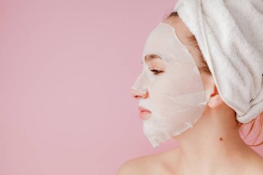 Güzel genç kadın kozmetik doku maske pembe bir arka plan üzerinde bir yüzünde uyguluyor. Sağlık ve güzellik tedavi ve teknoloji kavramı