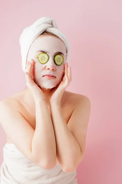 Mulher bonita está aplicando uma máscara de tecido cosmético em um rosto com pepino em um fundo rosa — Fotografia de Stock