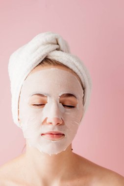 Güzel genç kadın kozmetik doku maske pembe bir arka plan üzerinde bir yüzünde uyguluyor. Sağlık ve güzellik tedavi ve teknoloji kavramı