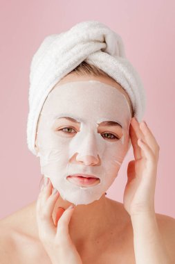 Güzel genç kadın kozmetik doku maske pembe bir arka plan üzerinde bir yüzünde uyguluyor. Sağlık ve güzellik tedavi ve teknoloji kavramı