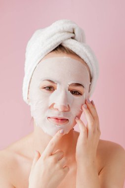 Güzel genç kadın kozmetik doku maske pembe bir arka plan üzerinde bir yüzünde uyguluyor. Sağlık ve güzellik tedavi ve teknoloji kavramı