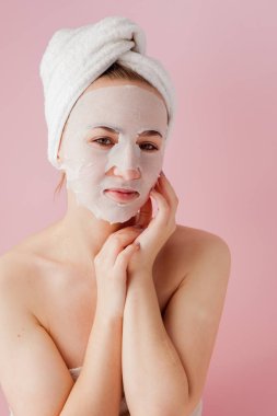 Güzel genç kadın kozmetik doku maske pembe bir arka plan üzerinde bir yüzünde uyguluyor