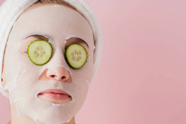 Wanita muda yang cantik menerapkan masker jaringan kosmetik pada wajah dengan mentimun pada latar belakang merah muda — Stok Foto