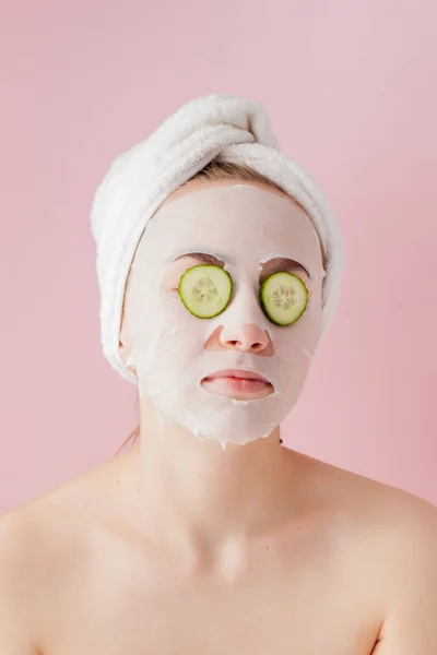 Mulher bonita está aplicando uma máscara de tecido cosmético em um rosto com pepino em um fundo rosa — Fotografia de Stock