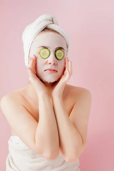 Mulher bonita está aplicando uma máscara de tecido cosmético em um rosto com pepino em um fundo rosa — Fotografia de Stock