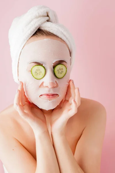 Mulher bonita está aplicando uma máscara de tecido cosmético em um rosto com pepino em um fundo rosa — Fotografia de Stock