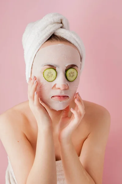 Krásná mladá žena je použití kosmetické tkáňové masky na tváři s okurkou na růžovém pozadí — Stock fotografie
