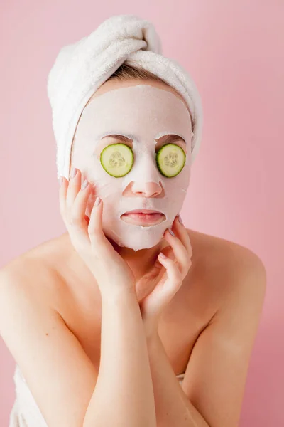 Mulher bonita está aplicando uma máscara de tecido cosmético em um rosto com pepino em um fundo rosa — Fotografia de Stock