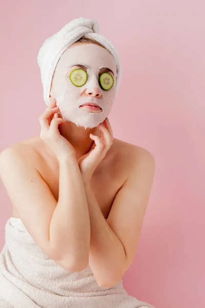 Hermosa mujer joven está aplicando una máscara de tejido cosmético en una cara con pepino sobre un fondo rosa — Foto de Stock