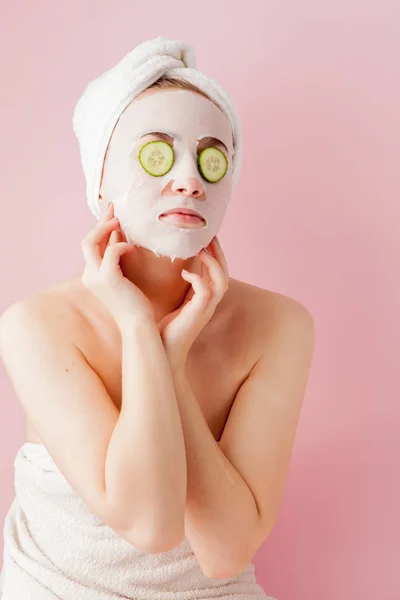 Mooie jonge vrouw is het aanbrengen van een cosmetische weefsel masker op een gezicht met komkommer op een roze achtergrond — Stockfoto