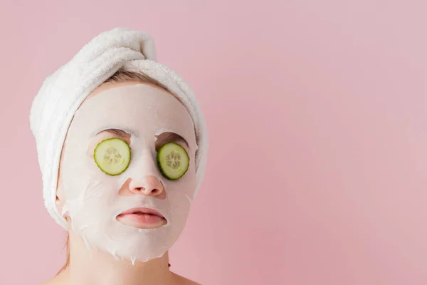 Mooie jonge vrouw is het aanbrengen van een cosmetische weefsel masker op een gezicht met komkommer op een roze achtergrond — Stockfoto