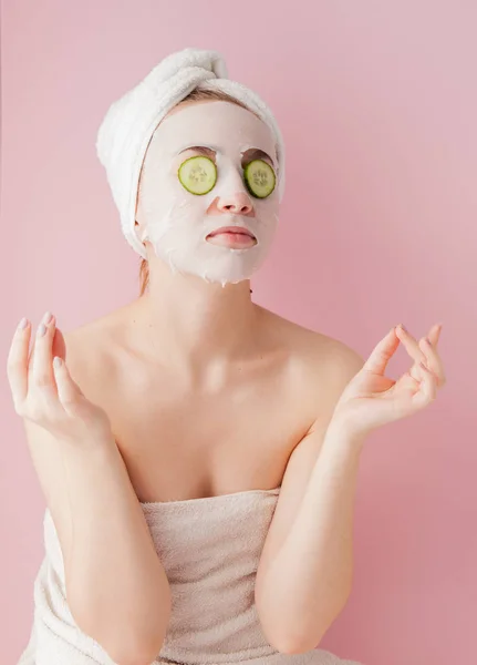 Wanita muda yang cantik menerapkan masker jaringan kosmetik pada wajah dengan mentimun pada latar belakang merah muda — Stok Foto