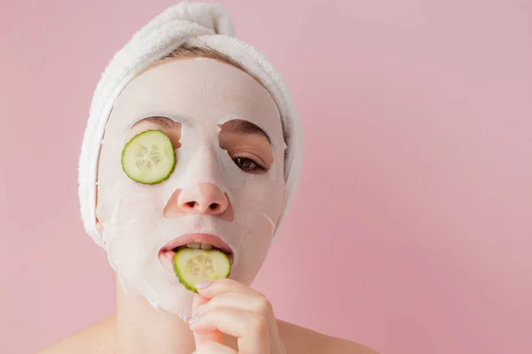 Wanita muda yang cantik menerapkan masker jaringan kosmetik pada wajah dengan mentimun pada latar belakang merah muda — Stok Foto