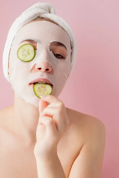 Mulher bonita está aplicando uma máscara de tecido cosmético em um rosto com pepino em um fundo rosa — Fotografia de Stock