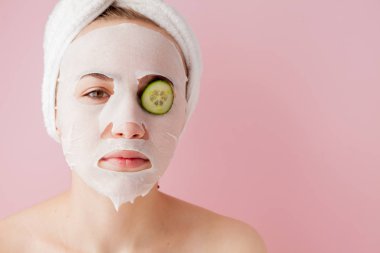 Güzel genç kadın kozmetik doku maske salatalığın üzerinde pembe bir arka plan ile bir yüzünde uyguluyor. Sağlık ve güzellik tedavi ve teknoloji kavramı