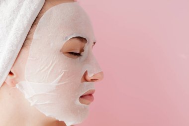 Güzel genç kadın kozmetik doku maske pembe bir arka plan üzerinde bir yüzünde uyguluyor. Sağlık ve güzellik tedavi ve teknoloji kavramı
