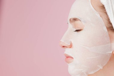 Güzel genç kadın kozmetik doku maske pembe bir arka plan üzerinde bir yüzünde uyguluyor. Sağlık ve güzellik tedavi ve teknoloji kavramı