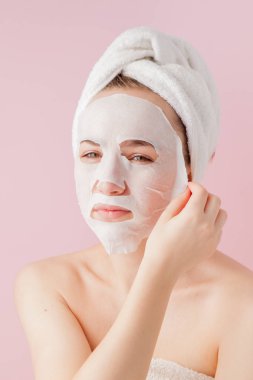 Güzel genç kadın kozmetik doku maske pembe bir arka plan üzerinde bir yüzünde uyguluyor. Sağlık ve güzellik tedavi ve teknoloji kavramı