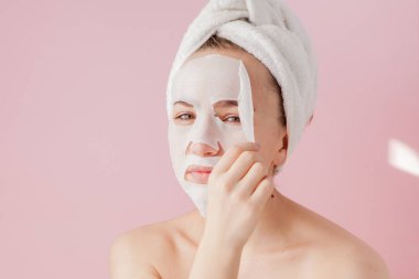 Güzel genç kadın kozmetik doku maske pembe bir arka plan üzerinde bir yüzünde uyguluyor. Sağlık ve güzellik tedavi ve teknoloji kavramı