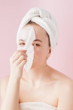Güzel genç kadın kozmetik doku maske pembe bir arka plan üzerinde bir yüzünde uyguluyor. Sağlık ve güzellik tedavi ve teknoloji kavramı