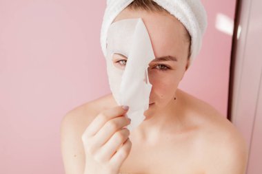 Güzel genç kadın kozmetik doku maske pembe bir arka plan üzerinde bir yüzünde uyguluyor. Sağlık ve güzellik tedavi ve teknoloji kavramı