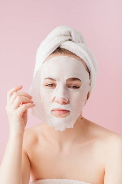 Güzel genç kadın kozmetik doku maske pembe bir arka plan üzerinde bir yüzünde uyguluyor. Sağlık ve güzellik tedavi ve teknoloji kavramı