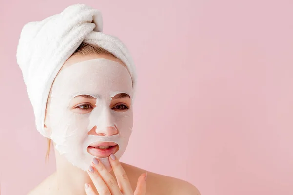 Hermosa mujer joven está aplicando una máscara de tejido cosmético en una cara sobre un fondo rosa — Foto de Stock