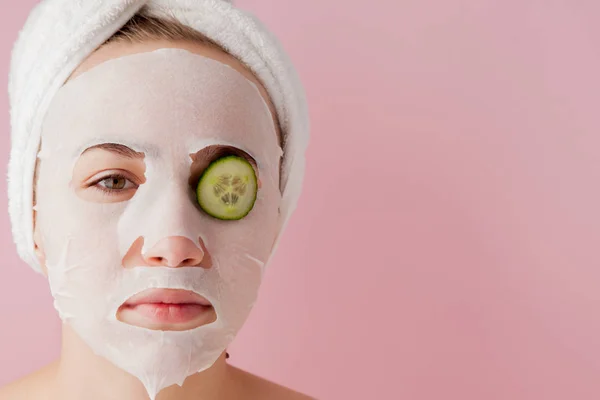 Wanita muda yang cantik menerapkan masker jaringan kosmetik pada wajah dengan mentimun pada latar belakang merah muda. Kesehatan dan kecantikan pengobatan dan konsep teknologi — Stok Foto
