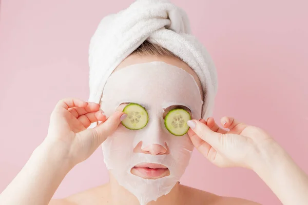 Wanita muda yang cantik menerapkan masker jaringan kosmetik pada wajah dengan mentimun pada latar belakang merah muda. Kesehatan dan kecantikan pengobatan dan konsep teknologi — Stok Foto