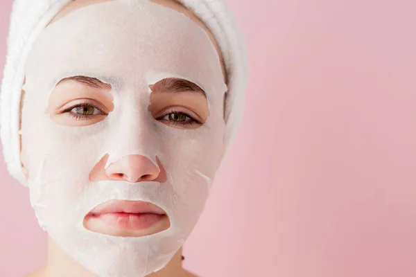 Kaunis nuori nainen soveltaa kosmeettinen kudos naamio kasvot vaaleanpunainen tausta. Terveydenhuollon ja kauneudenhoidon ja teknologian käsite — kuvapankkivalokuva