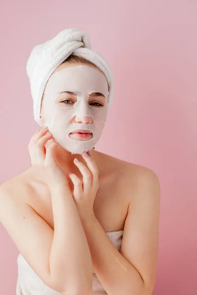 Gyönyörű fiatal nő az alkalmazása a kozmetikai szövet maszk egy arc, egy rózsaszín háttér. Egészségügyi- és szépségápolási kezelés és a technológia koncepció — Stock Fotó