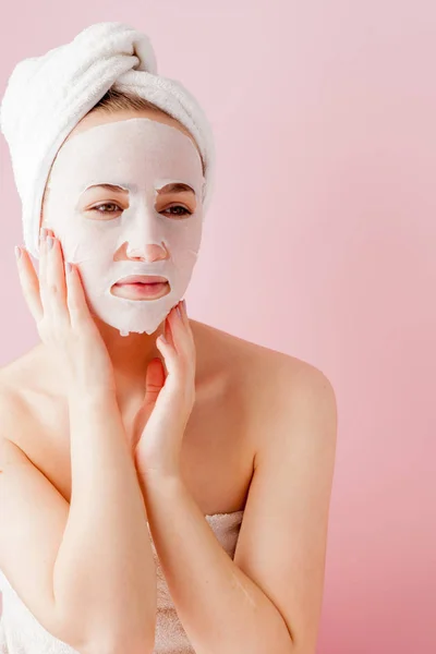 Hermosa mujer joven está aplicando una máscara de tejido cosmético en una cara sobre un fondo rosa. Salud y belleza tratamiento y concepto de tecnología — Foto de Stock