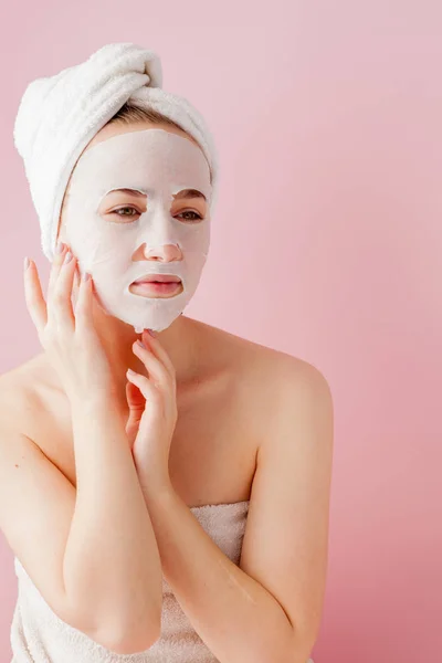 Kaunis nuori nainen soveltaa kosmeettinen kudos naamio kasvot vaaleanpunainen tausta. Terveydenhuollon ja kauneudenhoidon ja teknologian käsite — kuvapankkivalokuva