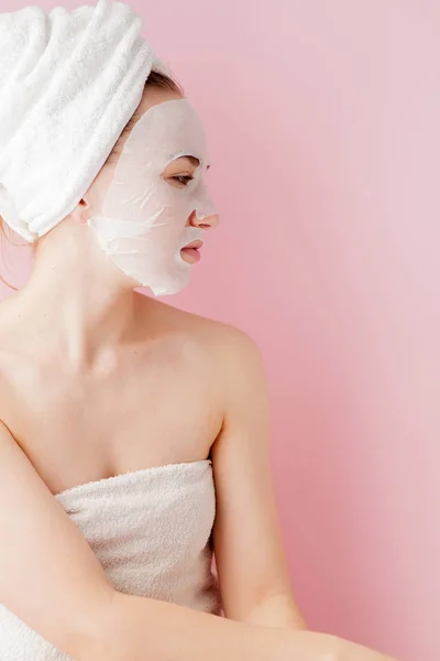 Hermosa mujer joven está aplicando una máscara de tejido cosmético en una cara sobre un fondo rosa. Salud y belleza tratamiento y concepto de tecnología —  Fotos de Stock