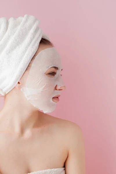 Wanita muda yang cantik menerapkan masker jaringan kosmetik pada wajah dengan latar belakang merah muda. Kesehatan dan kecantikan pengobatan dan konsep teknologi — Stok Foto