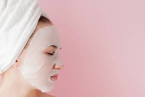 Wanita muda yang cantik menerapkan masker jaringan kosmetik pada wajah dengan latar belakang merah muda. Kesehatan dan kecantikan pengobatan dan konsep teknologi — Stok Foto