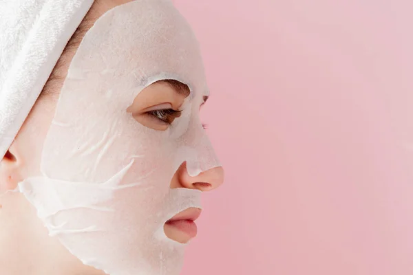 Wanita muda yang cantik menerapkan masker jaringan kosmetik pada wajah dengan latar belakang merah muda. Kesehatan dan kecantikan pengobatan dan konsep teknologi — Stok Foto