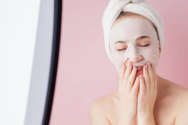 Hermosa mujer joven está aplicando una máscara de tejido cosmético en una cara sobre un fondo rosa. Salud y belleza tratamiento y concepto de tecnología — Foto de Stock