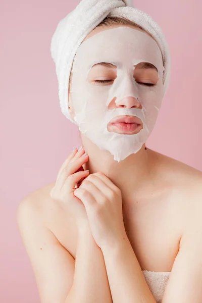 Güzel genç kadın kozmetik doku maske pembe bir arka plan üzerinde bir yüzünde uyguluyor. Sağlık ve güzellik tedavi ve teknoloji kavramı