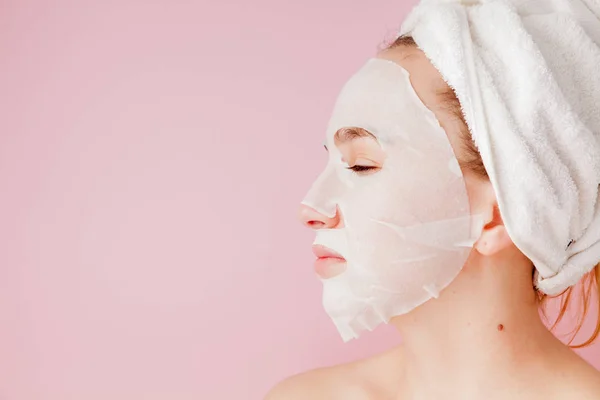 Hermosa mujer joven está aplicando una máscara de tejido cosmético en una cara sobre un fondo rosa. Salud y belleza tratamiento y concepto de tecnología — Foto de Stock