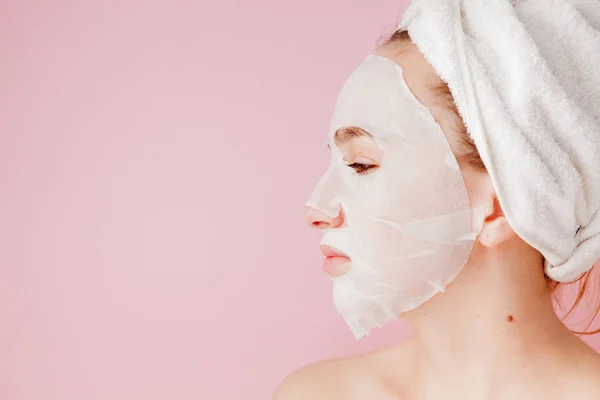 Belle jeune femme applique un masque de tissu cosmétique sur un visage sur un fond rose. Soins de santé et soins de beauté et concept technologique — Photo