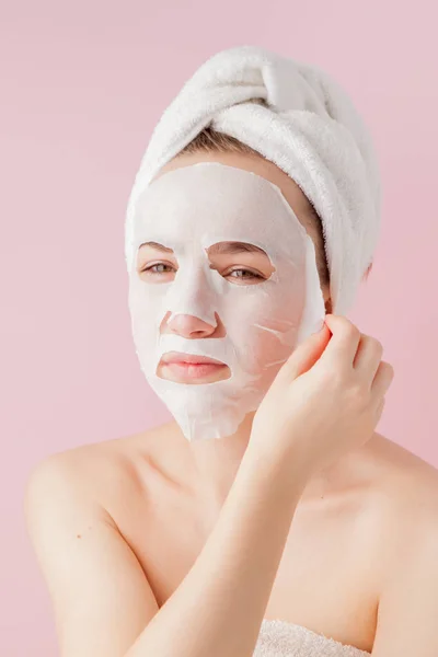 Hermosa mujer joven está aplicando una máscara de tejido cosmético en una cara sobre un fondo rosa. Salud y belleza tratamiento y concepto de tecnología — Foto de Stock