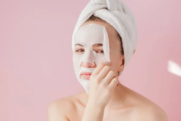 Wanita muda yang cantik menerapkan masker jaringan kosmetik pada wajah dengan latar belakang merah muda. Kesehatan dan kecantikan pengobatan dan konsep teknologi — Stok Foto