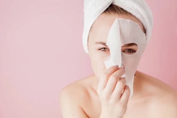 Όμορφη νεαρή γυναίκα εφαρμόζει μια μάσκα καλλυντικών ιστού σε ένα πρόσωπο σε ροζ φόντο. Υγείας και ομορφιάς θεραπείας και τεχνολογία έννοια — Φωτογραφία Αρχείου
