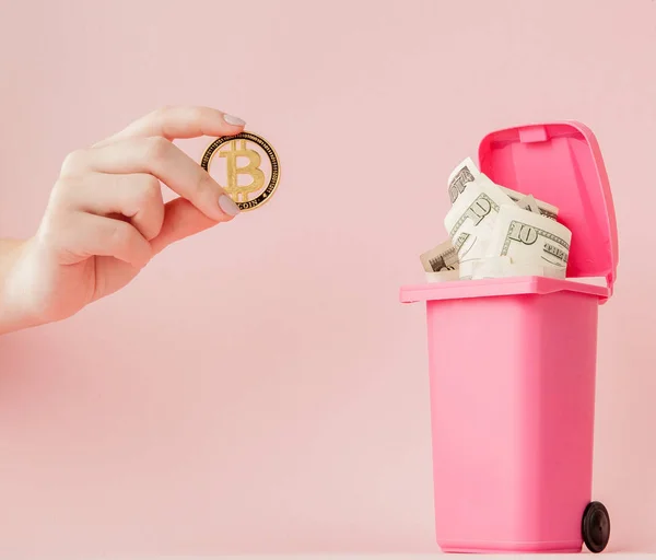 Bitcoin na mão de uma mulher e dólares em uma lata de lixo no fundo rosa — Fotografia de Stock