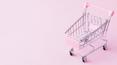 Küçük süpermarket bakkal itme sepeti oyuncak alışveriş için tekerlekler ve pembe plastik elemanları pembe pastel renkli kağıda düz arka plan yattı. Alışveriş kavramı. Kopya alanı reklam için