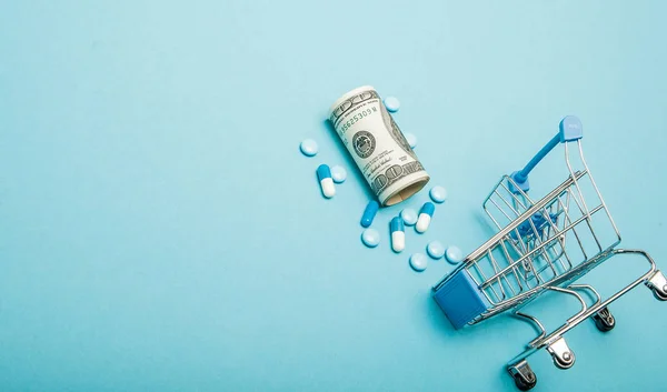 Dollars, pillen en winkelwagen op een blauwe achtergrond. Farmaceutisch concept. Kopieerruimte — Stockfoto