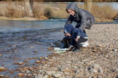 Kaydet çevre kavramı, küçük bir çocuk ve annesi çöp içine çöpe atılmış plaj üzerinde atık ve plastik şişeler topluyor.