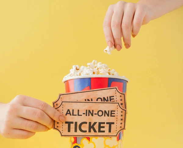 Hand tar en biobiljett och popcorn från en papperskopp på en gul bakgrund. Kvinnan äter popcorn. Filmkoncept. Ett platt ligg. Kopiera utrymme — Stockfoto