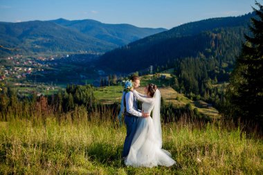 Yeni evliler gülümseme ve diğer her dağın zirvesinde çayır arasında sarıl. Düğün yürüyüş dağlar, nazik duygular çift, Sevgililer günü için fotoğraf ormanda